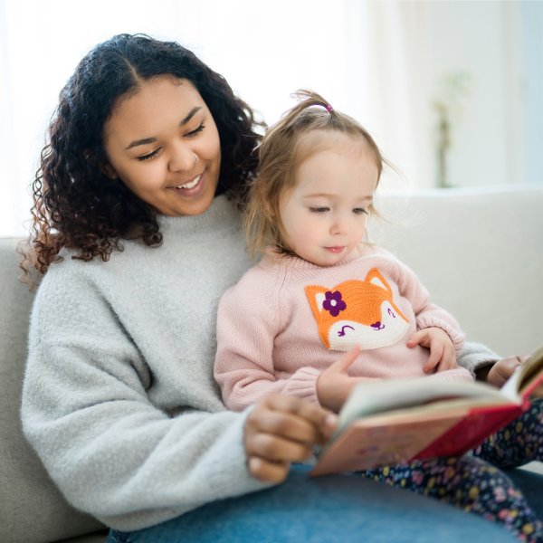 A young Black female babysitter and a little blonde girl sitting on a couch and reading a book together