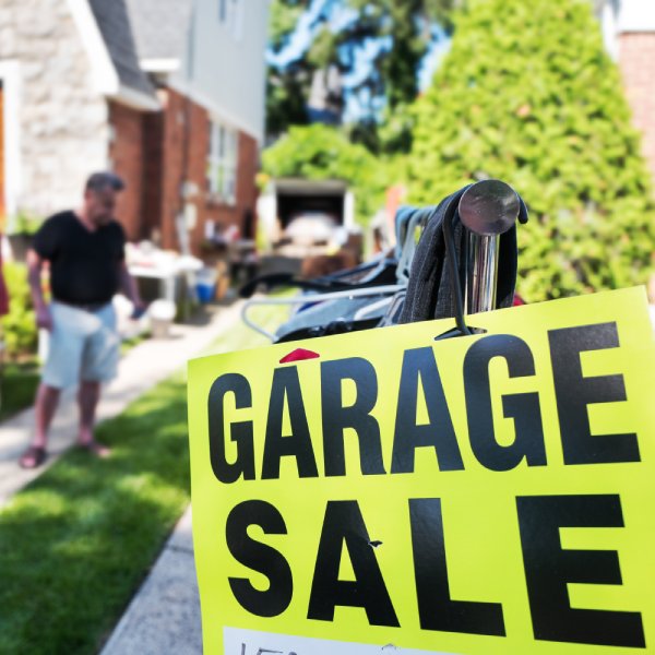 A garage sale sign in front of a house with people browsing items on blankets on the lawn in the background.