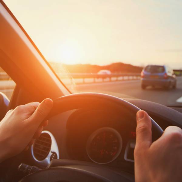 A pair of hands grip the steering wheel of a car that is driving in the right lane of a highway at sunset.